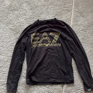 Svart långärmad tröja EA7 Armani - Svart långärmad tröja från EA7 Emporio Armani med stort guldtryck på bröstet. Snygg och stilren design med rund hals och mjukt bomullsmaterial. Perfekt för dig som gillar exklusiva märken och vill sticka ut med en fet logga. Xs men passar S