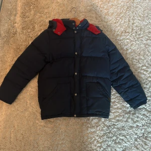Mörkblå pufferjacka Polo Ralph Lauren - Snygg mörkblå pufferjacka från Polo Ralph Lauren med röda detaljer på huvan och insidan. Jackan har stora fickor, knappar med logga och broderad häst på bröstet. Perfekt för kalla dagar med sin vadderade design och klassiska look.