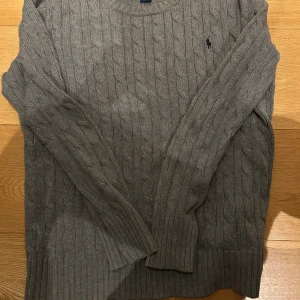 Grå kabelstickad tröja Polo Ralph Lauren - Kabelstickad grå tröja från Polo Ralph Lauren med klassisk rund hals och broderad marinblå logga på bröstet. Tröjan har långa ärmar och ribbade muddar för en snygg passform. Perfekt för lager-på-lager under kyliga dagar.
