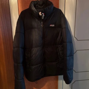 Svart pufferjacka från FILA - Svart pufferjacka från FILA med hög krage och diskret logga på bröstet. Jackan har dragkedja framtill och är quiltad för extra värme. Perfekt för kalla dagar och har en stilren look som funkar till det mesta.