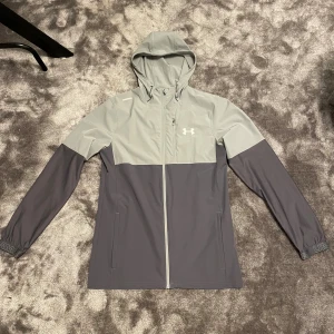 Grå vindjacka från Under Armour - Snygg tvåtonad grå vindjacka från Under Armour med huva och hel dragkedja framtill. Jackan har lång ärm, diskret logga på bröstet och praktiska fickor. Perfekt för träning eller när du vill ha en sportig look. Materialet är lätt och syntetiskt.