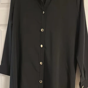 Svart set långskjorta med byxor från Brova Fashion  - Stilren svart set långskjorta och byxor från Brova Fashion med snygga knappar framtill. Skjortan har långa ärmar och en klassisk krage och byxan är lite vida. Perfekt för dig som gillar minimalistisk och tidlös stil. Storleken passar mer till 42. 