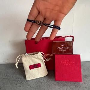 Valentino Garavani blå armband - Snyggt armband från Valentino Garavani med marinblå band och en silverfärgad metall-logga i mitten. Kommer med originalförpackning, tygpåse och certifikat. Perfekt accessoar för dig som gillar stilrena designer och exklusiva märken. (OBS KARTONGER OCH DUSTBAG INGÅR)(PRISET ÄR INRE HACKAT I STEN) (HAR KVITTO)