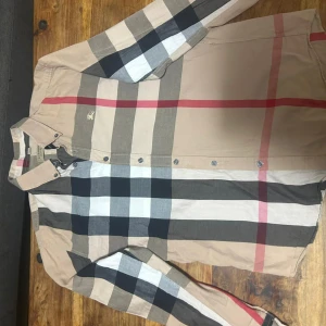 Rutig Burberry skjorta i beige och svart - Säljer en klassisk rutig skjorta från Burberry i storlek L men passar M. Skjortan har beige bas med svarta, vita, grå och röda rutor, knappar framtill och lång ärm. Ikonisk Burberry-logga broderad på bröstet. Tillverkad i mjuk bomull för en skön känsla.