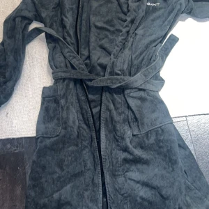 Mörkgrå badrock från GANT unisex - Mysig mörkgrå badrock från GANT i unisexmodell. Tillverkad i mjuk frotté med broderad logga på bröstet och knytskärp i midjan. Perfekt att slänga på sig efter duschen eller när du bara vill chilla hemma.
