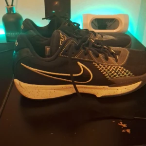 Svarta Nike sneakers med vit sula - Säljer ett par svarta Nike sneakers med vit, stänkmönstrad sula och gröna detaljer undertill. Skorna har snörning, tydlig Nike-logga på sidan och coolt zigzag-mönster i vitt och grönt. Perfekta för dig som gillar sportig stil och vill sticka ut. Använd några gånger förut