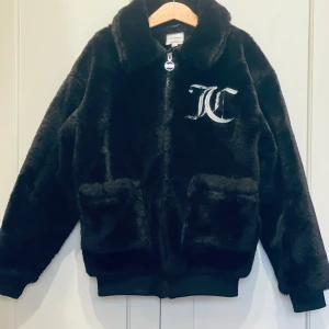 Svart pälsjacka från Juicy Couture - Mjuk och fluffig svart pälsjacka från Juicy Couture med stor broderad JC-logga på bröstet. Jackan har dragkedja med Juicy-detalj, två fickor framtill och klassisk krage. Perfekt för dig som vill ha en statementjacka med lyxig känsla.