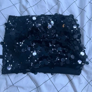 Svart paljettkjol i kort modell - Supersnygg svart kort kjol täckt med stora paljetter som glittrar i ljuset. Kjolen har en tight passform och är perfekt för dig som vill sticka ut. Materialet är mjukt och stretchigt, vilket gör den bekväm att bära hela kvällen.