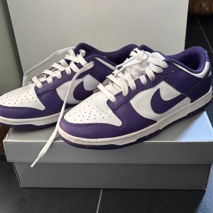 Nike Dunk Low vit/lila sneakers - Nike Dunk Low sneakers i vitt och lila skinn med klassisk swoosh på sidorna. Lågt skaft, vit snörning och vadderad plös med lila Nike-logga. Yttersulan är lila och ovandelen har perforerade detaljer för extra stil. Perfekta för dig som vill sticka ut med färg. Hjälper min brosa att sälja dessa skorna:) pris kan diskuteras vid snabbt köp!