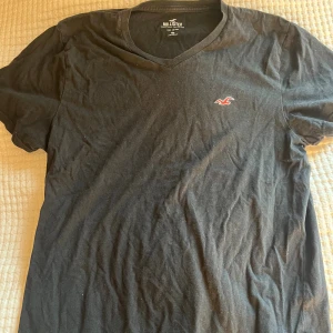 Svart v-ringad t-shirt från Hollister - Svart t-shirt från Hollister med v-ringning och liten röd logga broderad på bröstet. perfekt för en chill och enkel stil. Klassisk passform och korta ärmar gör den lätt att matcha med allt.