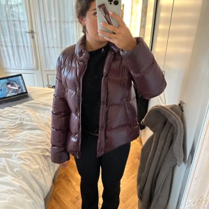 Mörkröd pufferjacka från Moncler - Säljer en snygg vinröd pufferjacka från Moncler med glansig finish. Jackan har hög krage, dragkedja med Moncler-detalj, två fickor med dragkedja och diskret logga på ärmen. Perfekt för kalla dagar och riktigt skön att bära.