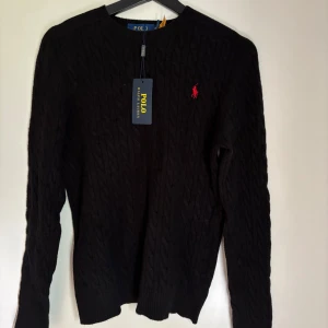 Svart stickad Polo Ralph lauren - Kabelstickad svart tröja från Polo Ralph Lauren med rund halsringning och röd broderad logga på bröstet. Klassisk och stilren design med långa ärmar och ribbade muddar. Storlek är M men den är lite tajt, aldrig använd.
