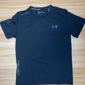 Svart Under Armour t-shirt - Snygg svart t-shirt från Under Armour med korta ärmar och coolt regnbågsskiftande logga på bröstet. Materialet är lätt och andas, perfekt för träning. Diskreta detaljer med text på axel och nederkant ger en sportig vibe.                              Finns i storlek M & L