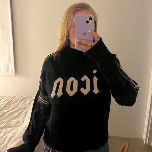 Svart stickad tröja Hunkydory 'icon' - Svart stickad tröja från Hunkydory med vit 'icon'-text framtill. Tröjan har ribbad hals, muddar och nederkant samt coola ränder i grönt, vitt och lila längs ärmarna. Perfekt för dig som vill ha en statement-piece i garderoben.