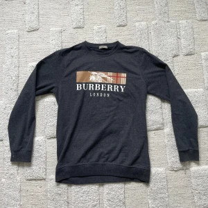 Mörkgrå Burberry sweatshirt  - Snygg mörkgrå sweatshirt från Burberry London i storlek XXL. Storlek skulle jag säga är som en M. Tröjan har ett klassiskt Burberry-tryck framtill i beige, rött och vitt med logga och rutor. Mjuk bomullskänsla och rund hals. Perfekt för dig som gillar stilrena och ikoniska plagg.