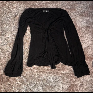 Svart långärmad topp med knyt från Design by Si - Svart långärmad topp från Design by Si med snygg knytning framtill. Toppens material är mjukt och stretchigt, perfekt för en trendig look. Den har en smickrande passform och lätt utsvängda ärmar som ger en cool vibe.