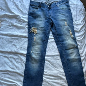 Blå jeans med slitningar och paljettdetalj - Snygga blå jeans från Panos Emporio med slitna detaljer och coola guldpaljetter under trasorna på benen. Klassisk femficksmodell i denim med raka ben och normal passform. Perfekta för dig som gillar stockholmsstil.