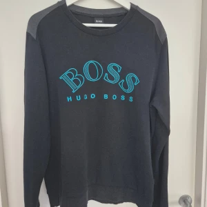 Svart sweatshirt från Hugo Boss - Svart sweatshirt från Hugo Boss med blått broderat BOSS-tryck på bröstet. Tröjan har grå axelpartier, rund halsringning och långa ärmar. Perfekt för en stilren och sportig look.