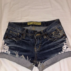 Jeansshorts från Gojin med brodyr - Snygga mörkblå jeansshorts från Gojin med broderade detaljer på bakfickorna och vit spets vid bensluten. Shortsen har låg midja, fransiga kanter och klassisk femficksmodell. Perfekta för sommaren och ger en cool, avslappnad vibe.