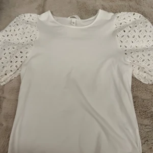 Vit blus med broderade puffärmar - Superfin vit blus från H&M med broderade puffärmar i spets och rund halsringning. Ärmarna är korta och har ett snyggt hålmönster som ger en romantisk vibe. Perfekt att styla med jeans eller kjol för en fräsch look. Aldrig andvänd!!!!