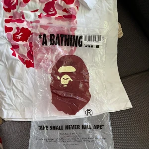 Vit BAPE t-shirt  - Säljer en vit t-shirt från A Bathing Ape med ikoniskt camo-apa tryck i rött och rosa på bröstet. Klassisk passform med korta ärmar och crewneck. Materialet är mjuk bomull och det finns en liten BAPE-logga på ärmen. Perfekt för dig som gillar streetwear och vill sticka ut.