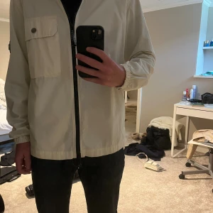 Beige vindjacka från Lagency - Snygg beige vindjacka från Lagency med svart logopatch på ärmen och en stor bröstficka med knapp. Jackan har dragkedja framtill, klassisk krage och elastiska muddar vid ärmsluten. Perfekt för dig som gillar stilren och clean look.