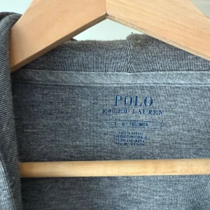 Grå hoodie från Polo Ralph Lauren - Snygg grå hoodie från Polo Ralph Lauren med dragkedja, huva med snörning och två stora fickor framtill. Klassisk logga broderad på bröstet. Perfekt för en chill och stilren look. Tillverkad i mjuk bomullsblandning för extra komfort.