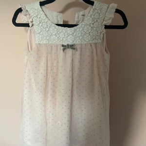 Söt prickig blus med spets och rosett 💘 - Supersöt ljusrosa blus med små guldprickar, volangdetaljer vid axlarna och bred spets upptill. Framtill sitter en liten grå rosett. Blusen är ärmlös och har en luftig, flowy passform. Perfekt för dig som gillar romantisk och feminin stil. Har tyvär ett hål som amn ser på andra bilden men syns inte supermycket 💘 vet inte riktigt storleken men är ganska liten runt armarna 🩷🤷‍♀️