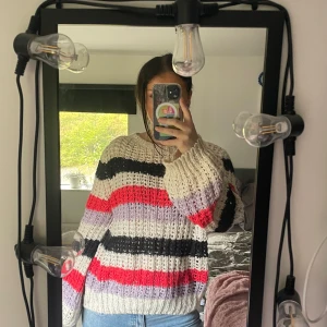 Randig stickad tröja i pastell och rött - Supermysig stickad tröja med breda ränder i vitt, svart, lila och rött. Tröjan har en loose passform och långa ärmar, perfekt för kyliga dagar. Den ribbade strukturen ger en cool och avslappnad vibe. Passar perfekt till jeans eller kjol.