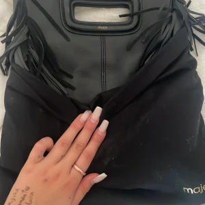 Svart fransad handväska från Maje - Säljer en svart handväska från Maje i skinn med coola fransdetaljer på sidorna och ett inbyggt handtag. Väskan har en stilren, fyrkantig form och diskret logga i guld vid handtaget. Perfekt för dig som vill ha en statementväska med edge.