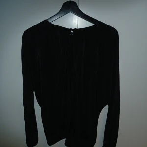 Svart långärmad topp med öppen rygg - Säljer en svart långärmad topp i glansigt, plisserat tyg med snygg öppen rygg och knyt upptill. Toppen har en cool struktur och passar perfekt när du vill sticka ut lite extra. Perfekt för dig som gillar trendiga detaljer och en unik look.