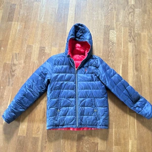 Vändbar pufferjacka Polo Ralph Lauren - Vändbar pufferjacka från Polo Ralph Lauren i barnstorlek xl passar 160-170. Ena sidan är marinblå och andra sidan röd, båda med broderad logga på bröstet. Jackan har huva, dragkedja och två fickor med knappstängning. Perfekt för kalla dagar och enkel att matcha. Liten hål i ena armen vilket visas på sista bilden.