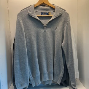 Grå half zip tröja Polo Ralph Lauren - Snygg grå långärmad tröja från Polo Ralph Lauren med half zip och klassisk krage. Tröjan är i bomull och har en diskret broderad logga på bröstet. Perfekt för dig som gillar stilren och enkel design.