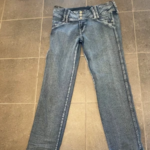 Blå jeans med paljettdetaljer - Snygga blå jeans med låg midja, raka ben och coola paljettdetaljer på linningen. Bälteshällorna är dekorerade med små diamanter och jeansen har tre dekorativa knappar framtill. Perfekt för dig som gillar extra detaljer på dina jeans.står ingen storlek men skulle säga small/ medium! Använda två gånger.