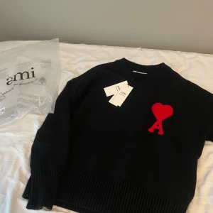 Svart stickad tröja från Ami - Svart stickad tröja från Ami Alexandre Mattiussi med röd hjärtformad A-logga på bröstet. Tröjan har rund hals, ribbade muddar och är långärmad. Materialet är mjuk ull som känns riktigt skönt mot huden. Perfekt för dig som gillar stilrena detaljer och snygg design.