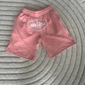 Rosa shorts från Corteiz - Säljer ett par snygga rosa shorts från Corteiz i storlek M. De har elastisk midja med vit snörning och ett vitt tryck framtill. Sköna och avslappnade med lite loose fit. Dom har fläckar på sig men det går att tvätta bort.