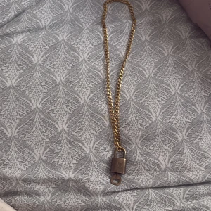 Louis Vuitton halsband med lås - säljer Unikt halsband från Louis Vuitton med grov guldfärgad kedja och hängande lås i metall. Låset har LV-loggan på framsidan och graverad text på baksidan. 