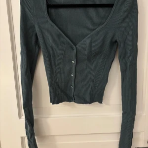 Ribbad mörkgrön topp från H&M XS - Säljer en ribbad mörkgrön topp från H&M i storlek XS. Toppen har v-ringning, långa ärmar och knappar framtill. Den är croppad och har en slim passform som sitter snyggt på kroppen. 