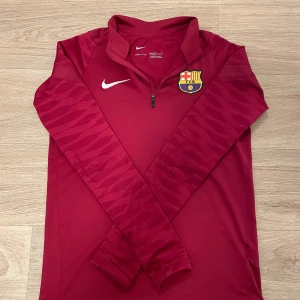 Barcelona långärmad träningströja Nike - Snygg vinröd långärmad fotbollströja från Nike med FC Barcelona-emblem på bröstet. Tröjan har halv dragkedja och diskret mönster på ärmarna. Tillverkad i funktionsmaterial som andas, perfekt för träning eller match.