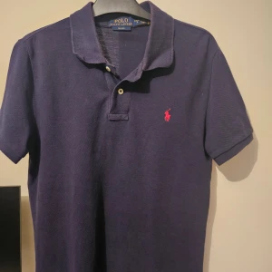 Mörkblå pikétröja från Polo Ralph Lauren - Säljer en klassisk mörkblå pikétröja från Polo Ralph Lauren med korta ärmar och rosa broderad logga på bröstet. Tröjan har krage och två knappar framtill. Tillverkad i bomull för en skön och luftig känsla.