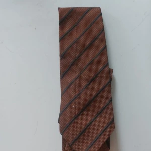 Brun randig slips från Portia - Snygg brun slips från Portia med diagonala svarta ränder och diskret rutmönster. Tillverkad i mikrofiber, vilket ger en mjuk och lätt känsla. Perfekt för dig som vill ha en klassisk men ändå modern look. Made in Sweden.