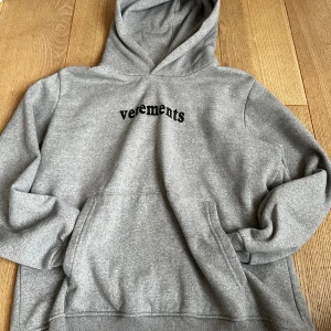 Grå hoodie från Vetements - Säljer en grå hoodie från Vetements med svart broderad logga på bröstet och coolt postetikett-tryck på ryggen. Hoodien har stor huva, magficka och mjukt fleecetyg på insidan. Perfekt för dig som gillar streetwear och vill sticka ut.