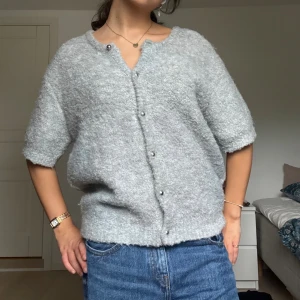 Ljusgrå stickad kofta med pärlknappar - Supermjuk kortärmad ljusgrå stickad kofta med snygga pärlknappar framtill. Koftan har en avslappnad passform och ribbade kanter vid ärmslut och nederkant. 
