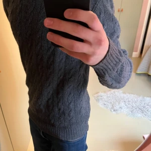 Mörkgrå stickad tröja Polo Ralph Lauren - Säljer en mörkgrå stickad tröja från Polo Ralph Lauren med klassiskt kabelstickat mönster. Tröjan har rund hals och långa ärmar, perfekt för dig som gillar stilren design. Passar dig som vill ha en mysig och snygg tröja till hösten och vintern.