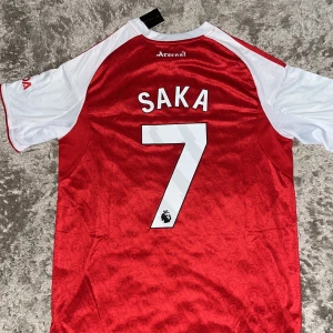 Arsenal Saka 7 matchtröja Adidas S - Arsenal hemmatröja med Saka 7 på ryggen. Röd kropp med vita ärmar, klassiska Adidas-ränder på axlarna och klubbmärke på bröstet. Tillverkad i lätt och ventilerande polyester med subtilt mönster i tyget. Slim fit-modell, officiellt licensierad produkt.
