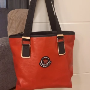 Röd axelväska från Moncler - Cool röd axelväska från Moncler med svart logotyp framtill. Väskan har breda svarta axelband med Moncler-tryck, svarta detaljer och dragkedja upptill. Tillverkad i syntetmaterial med rymligt fack och innerficka med dragkedja. Vattentålig! 