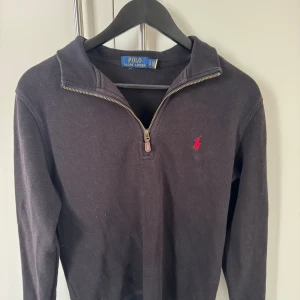 Svart half zip tröja från Polo Ralph Lauren - Svart långärmad tröja från Polo Ralph Lauren med half zip och klassisk krage. Röd broderad logga på bröstet ger en snygg kontrast. Tröjan är gjord i mjuk bomull och har en stilren, normal passform som funkar till många olika looks.