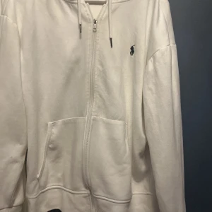 Vit hoodie från Ralph Lauren - Vit hoodie från Ralph Lauren med dragkedja framtill, huva med snörning och två stora fickor. Klassisk broderad logga på bröstet. Perfekt för en avslappnad och stilren look.