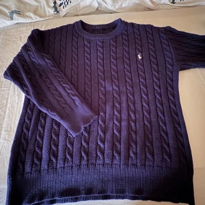 Mörklila stickad tröja från Ralph Lauren - Säljer en mörkblå/ marinblå kabelstickad tröja från Ralph Lauren med klassisk rund halsringning och broderad logga på bröstet. Tröjan har långa ärmar och ribbade muddar vid ärmslut och nederkant. Perfekt för lager-på-lager under kyliga dagar. Storlek- M/L den är stretchig 