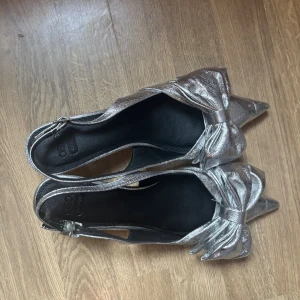 Silvriga klackar - Snygga silvriga pumps från ASOS med spetsig tå och rosett framtill. Har använt endast använt en gång. Klackarna är i storlek 39. Hör av för fler frågor❤️